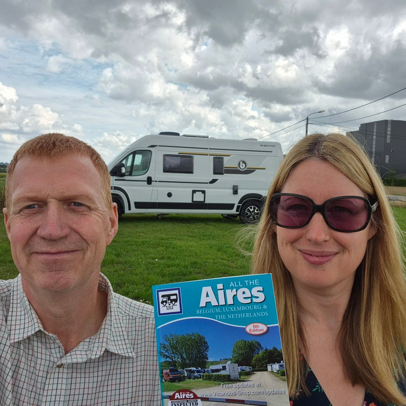 Chris and Meli at an aire in front of their motorhome holding All the Aires. located at grassy field under a cloudy sky.
